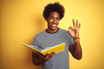 Afro american student man reading book standing over isolated yellow background doing ok sign with fingers, excellent symbol
