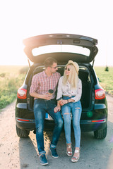 Young woman sitting in car trunk with her boyfriend having coffee. Couple on road trip.