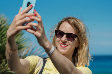 Young, beautiful woman takes selfie by sea on her phone smiling to camera. Happy girl takes photo on her smartphone on beach on sunny day.