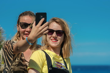  Young beautiful European girls in sunglasses taking selfie on cellphone at seacoast, during summer holidays.