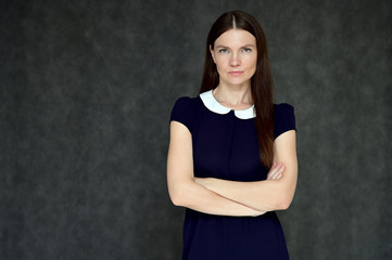 Portrait of a pretty young brunette woman with combed hair on a gray background in a dark blue dress. It is in different poses. Looks straight at the camera. Slim figure.