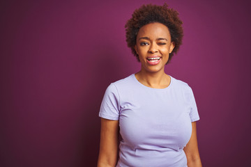 Obraz premium Young beautiful african american woman with afro hair over isolated purple background winking looking at the camera with sexy expression, cheerful and happy face.