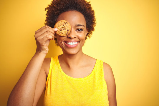 African american woman holding chocolate chips cookie over yellow background with a happy face standing and smiling with a confident smile showing teeth