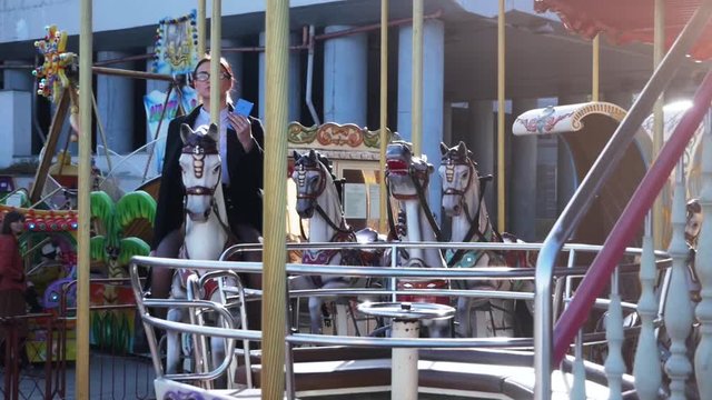 Funny scene. Young business woman is talking on the phone while riding a horse carousel. Office workers entertainment concept 4k