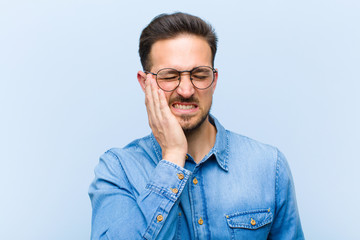 young handsome man holding cheek and suffering painful toothache, feeling ill, miserable and...