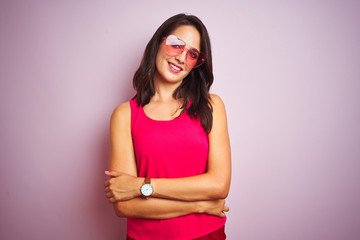 Beautiful woman wearing funny pink heart shaped sunglasses over pink isolated background happy face smiling with crossed arms looking at the camera. Positive person.