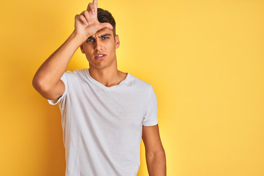 Young indian man wearing white t-shirt standing over isolated yellow background making fun of people with fingers on forehead doing loser gesture mocking and insulting.