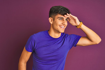 Fototapeta na wymiar Young indian man wearing t-shirt standing over isolated purple background very happy and smiling looking far away with hand over head. Searching concept.