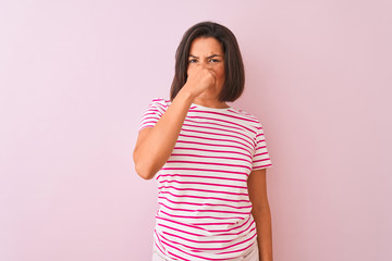 Young beautiful woman wearing striped t-shirt standing over isolated pink background smelling something stinky and disgusting, intolerable smell, holding breath with fingers on nose