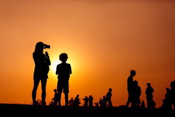 Fototapeta na wymiar silhouette of a woman holding a smartphone taking pictures with child and crowd people outside during sunset