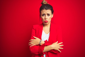 Young beautiful business woman standing over red isolated background shaking and freezing for winter cold with sad and shock expression on face