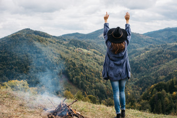 Rear view of young woman in hat stand near to campfire on top of mountain summit, raises arms into air, happy and drunk on life, youth and happiness. Explore travel real wilderness lifestyle