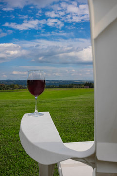 A glass of red wine photographed at on the arm rest of a white, plastic Adirondack chair near Cayuga Lake in the Finger Lakes Region of New York State.