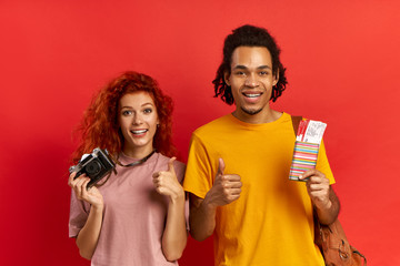 Happy triumphing diverse couple rejoices having trip soon, holds passport and flying tickets, showing thumbs up, holds old film camera and orange backpack, poses against red wall. Vacation concept