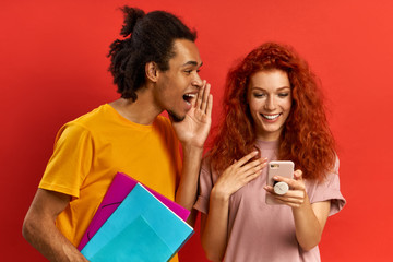Cheerful guy whispers secret to girlfriend shares gossip have fun after classes excited red haired female holds cell phone gets interesting information from groupmate. Two students discuss something