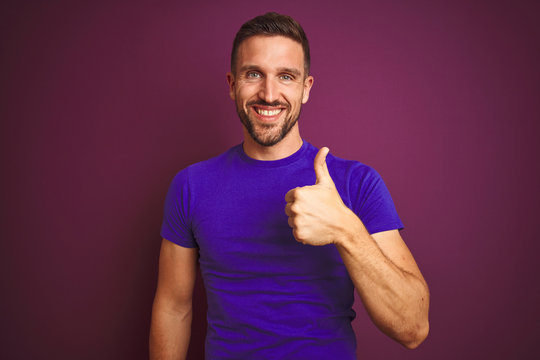 Young man wearing casual purple t-shirt over lilac isolated background doing happy thumbs up gesture with hand. Approving expression looking at the camera showing success.