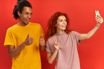 Happy dark skinned man makes okay gesture with both thumbs, poses at camera for making selfie together with girlfriend, she use modern cell phone shows victory or peace sign isolated on red background