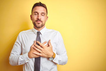 Young handsome business man wearing elegant white shirt over yellow isolated background smiling with hands on chest with closed eyes and grateful gesture on face. Health concept.