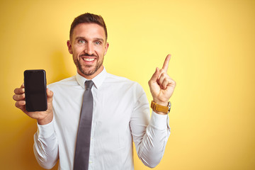 Young handsome business man showing smartphone screen over yellow isolated background very happy pointing with hand and finger to the side