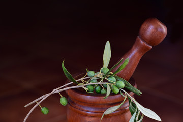 A mortar with wooden pestle against a tiled dark rustic background and an olive branch with green fresh olives on it