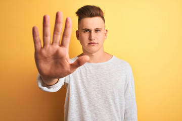 Young handsome man wearing white casual t-shirt standing over isolated yellow background doing stop sing with palm of the hand. Warning expression with negative and serious gesture on the face.
