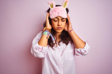 Young beautiful woman wearing pajama and sleep mask over isolated pink background with hand on head for pain in head because stress. Suffering migraine.