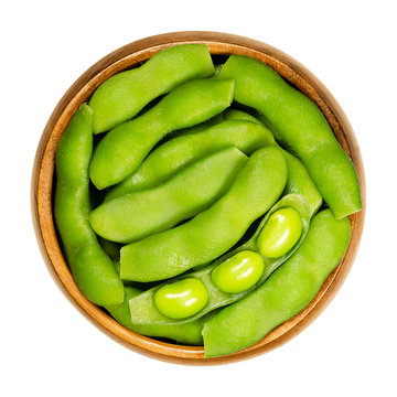 Green soybeans in the pod, edamame, in wooden bowl. Unripe soya beans, also Maodou. Glycine max, a legume, edible after cooking and rich protein source. Closeup, on white background, macro food photo.