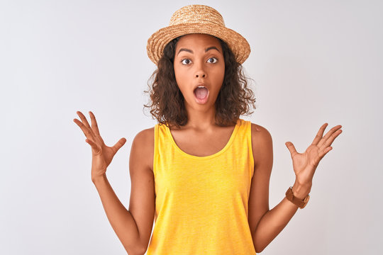 Young brazilian woman wearing yellow t-shirt and summer hat over isolated white background celebrating crazy and amazed for success with arms raised and open eyes screaming excited. Winner concept