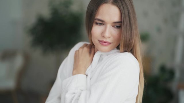 Portrait of young beautiful woman in white bathrobe looks into camera, smiles