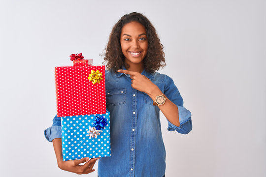 Young brazilian woman holding birthday gifts standing over isolated white background very happy pointing with hand and finger