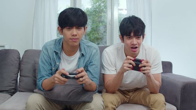 Young Asian gay couple play games at home, Teen korean LGBTQ men using joystick having funny happy moment together on sofa in living room at house.