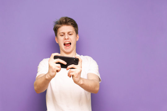 Expressive guy in a white T-shirt plays video games on a smartphone, looks at the screen and reacts emotionally. Young man plays a game on the phone, the focus is on a smartphone in his hands