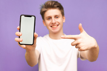 Smiling young man holds a modern smartphone in his hand and shows his finger on a blank white screen, isolated on a blue background. Focus on the smartphone in the hands of a happy guy.