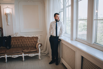 Stylish male businessman stands near the window. Wedding portrait of a handsome, bearded and smiling groom in the studio. Photography and concept.