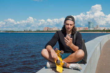 A young man sitting on a quay with a yellow skateboard on his lap is checking a smartphone.