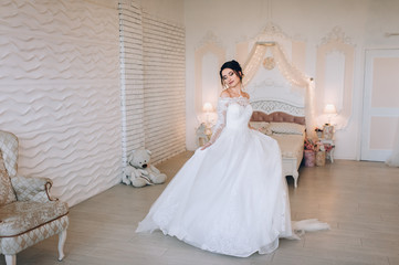 Fototapeta premium A beautiful bride is spinning and dancing, holding on to a dress. Wedding portrait of a cute girl in a room, studio on the background of wall and bed. Photography and concept. Wedding decorations.