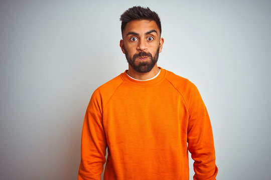 Young indian man wearing orange sweater over isolated white background puffing cheeks with funny face. Mouth inflated with air, crazy expression.