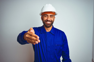 Handsome indian worker man wearing uniform and helmet over isolated white background smiling friendly offering handshake as greeting and welcoming. Successful business.