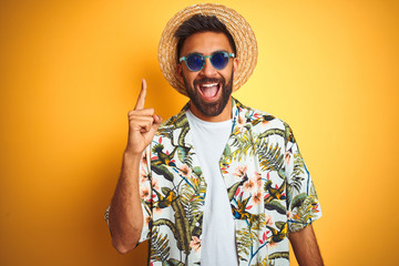 Indian man on vacation wearing floral shirt hat sunglasses over isolated yellow background pointing...