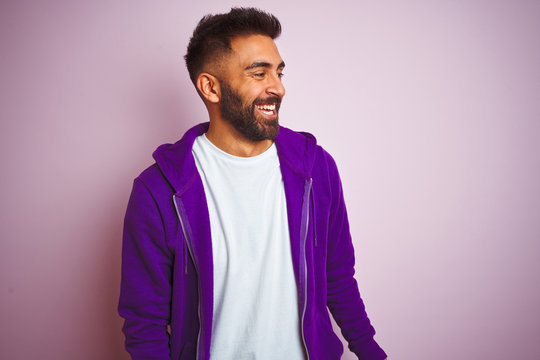 Young indian man wearing purple sweatshirt standing over isolated pink background looking away to side with smile on face, natural expression. Laughing confident.