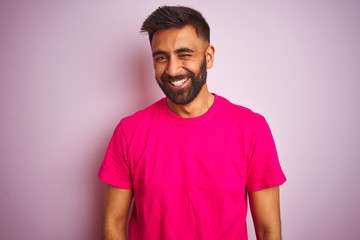 Young indian man wearing t-shirt standing over isolated pink background winking looking at the...