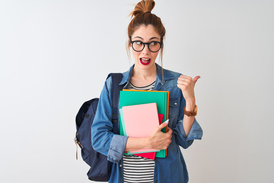 Redhead student woman wearing backpack holding books over isolated white background pointing and showing with thumb up to the side with happy face smiling