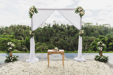 White Wedding ceremony arch in tropical jungle on edge of blue infinity swimming pool. Beautiful arch and decorations on the cliff.
