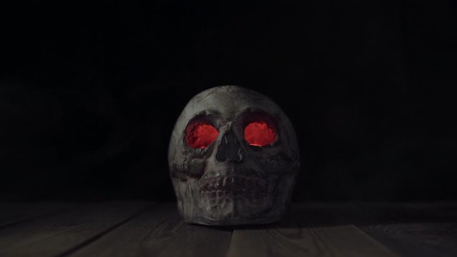 Old human skull with glowing eyes in smoke on a wooden table. Theme of human death and celebration of Halloween. Isolated death's head on a black background in a haze of fog.