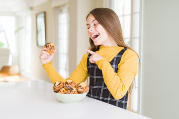 Beautiful young girl eating chocolate chips muffins very happy pointing with hand and finger