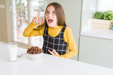 Beautiful young girl kid eating chocolate cereals and glass of milk for breakfast pointing finger...