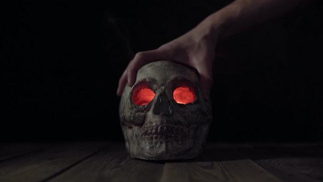 Man puts his hand on a human skull with glowing eyes and smoke coming out of them, then takes skull out of frame. Theme of human death. Isolated death's head on a black background in a haze of fog.