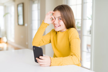 Beautiful young girl kid sending a message using smartphone with happy face smiling doing ok sign with hand on eye looking through fingers