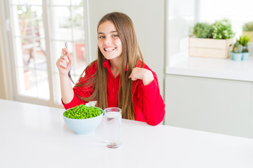Beautiful young girl eating healthy green peas with surprise face pointing finger to himself