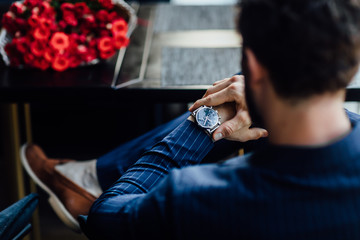 Photo of theback, businessman is sitting in modern restaurant with bouquet of red roses and checking the time while waiting for his beloved.